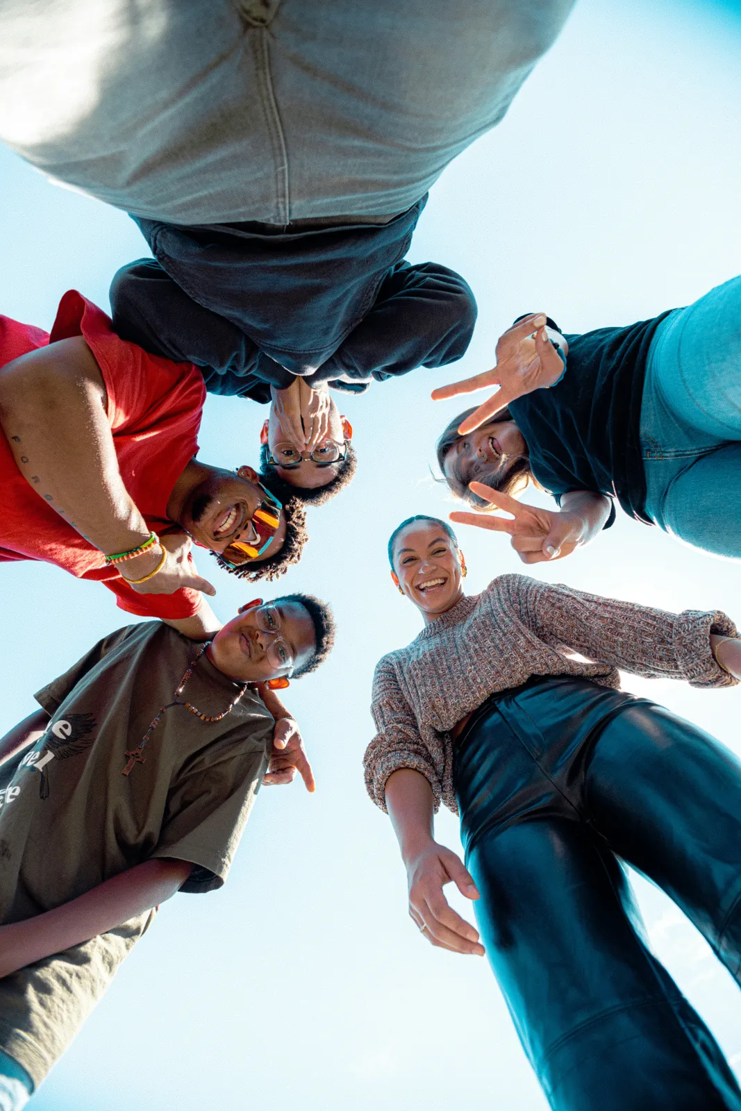 Photo: five friends standing in a circle, seen from below against a blue sky, laughing and making peace signs at the camera.