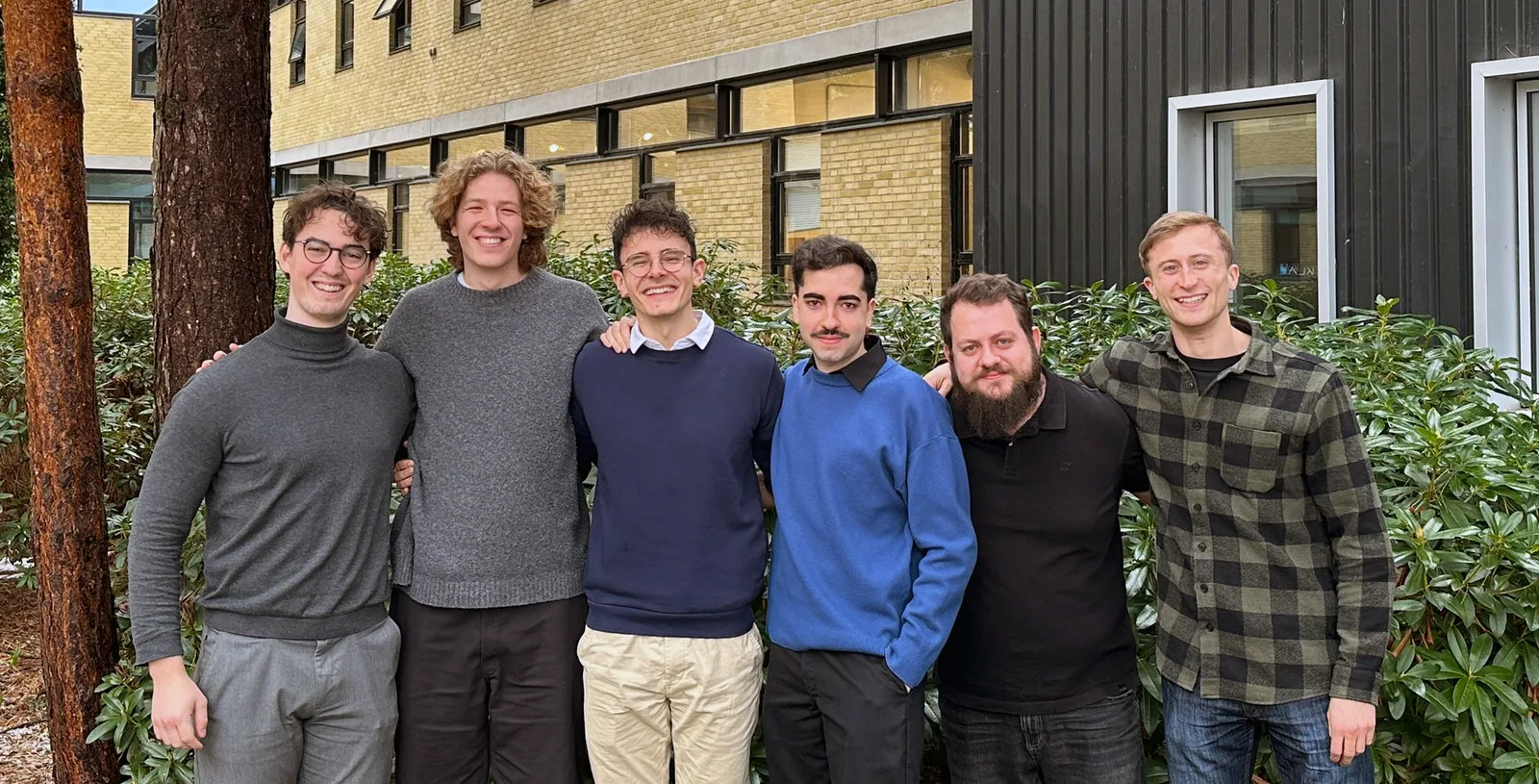 The six people behind Synoma standing shoulder to shoulder outdoors at DTU, smiling.